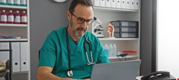 Hispanic man in hospital clinic room wearing medical scrubs and glasses