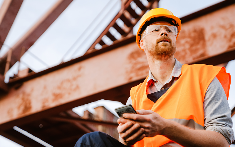 White ginger man wearing helmet and vest using cellphone working in port
